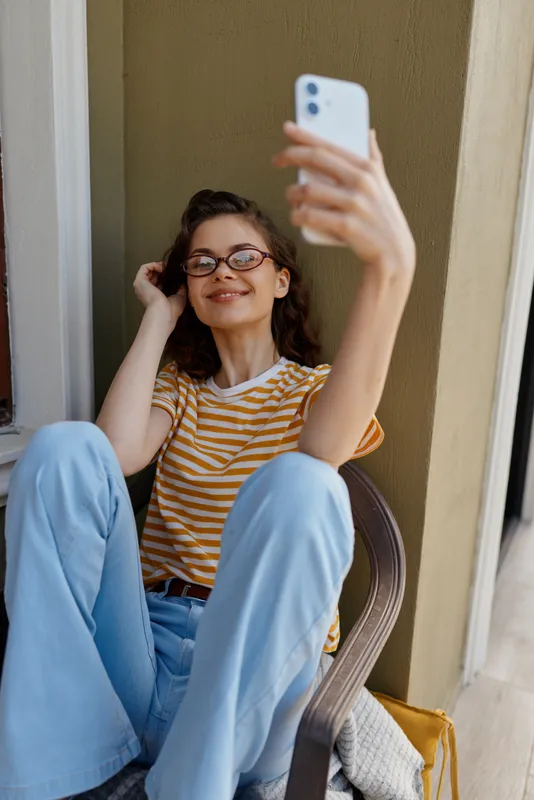 Young woman sits on the floor and takes a selfie with a smartphone, wearing striped shirt and jeans, cozy home setting, natural light, cheerful relaxed mood, casual lifestyle.