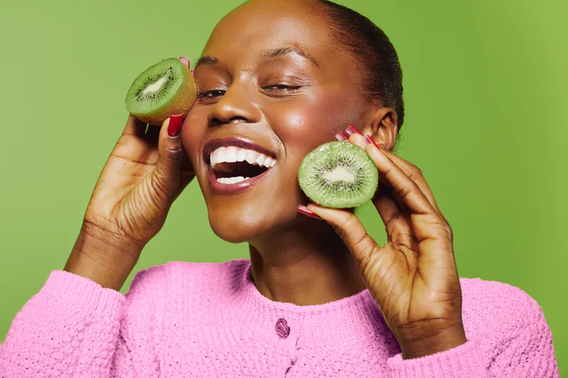 Happy African woman with dark skin holds two halves of fresh kiwi near face smiling joyfully on green background. Concept of healthy lifestyle, natural food, wellness and positive mood.