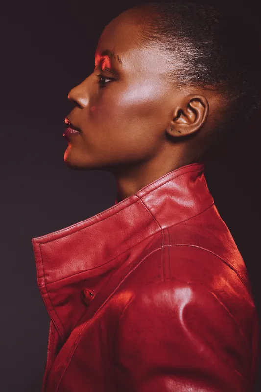 Profile of elegant black woman with dark skin wearing red leather jacket. Studio portrait on dark background. Concept of fashion, style, confidence, and beauty in modern culture.