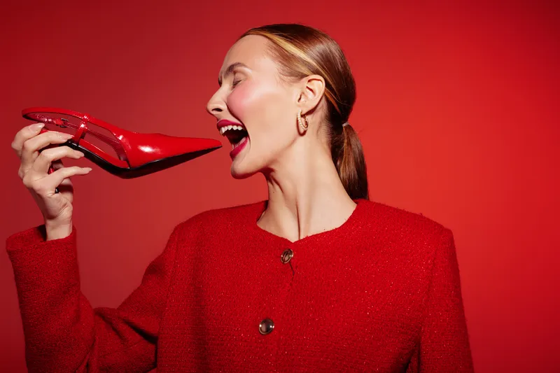 Woman in red jacket holds red high heel shoe near face with eyes closed and smiling. Studio portrait on red background. Concept of fashion, style, and joyful expression.