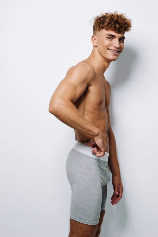 Young man with curly hair smiles and stands shirtless in gray shorts against a white wall, showing confidence and relaxed posture in casual lifestyle portrait.