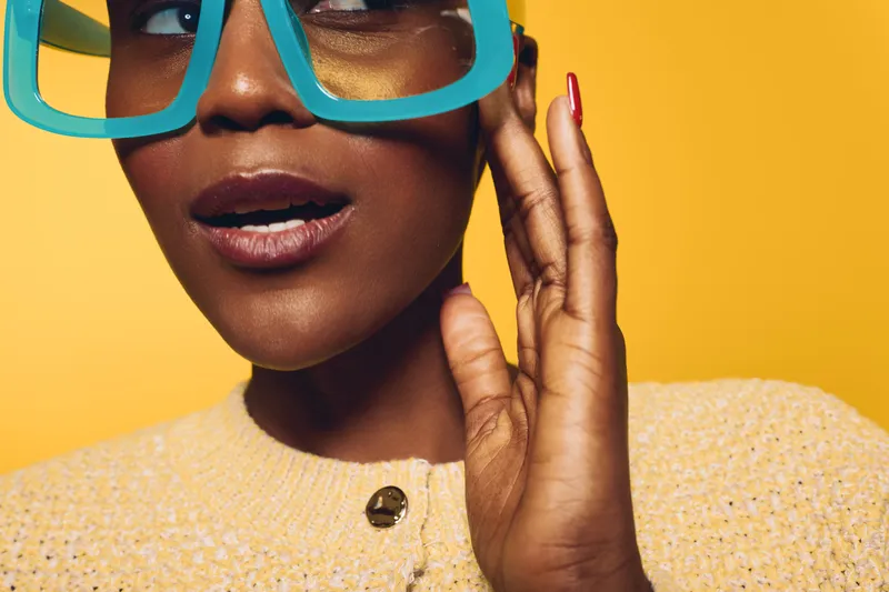 Close-up of young African woman with dark skin wearing oversized blue glasses and yellow sweater on bright yellow background. Concept of fashion, style, and beauty portrait.