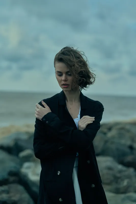 Moody coastal scene with a woman in a dark coat, arms crossed, gazing down as wind sweeps the sea and rocks. Dramatic, contemplative mood with subtle fashion appeal.
