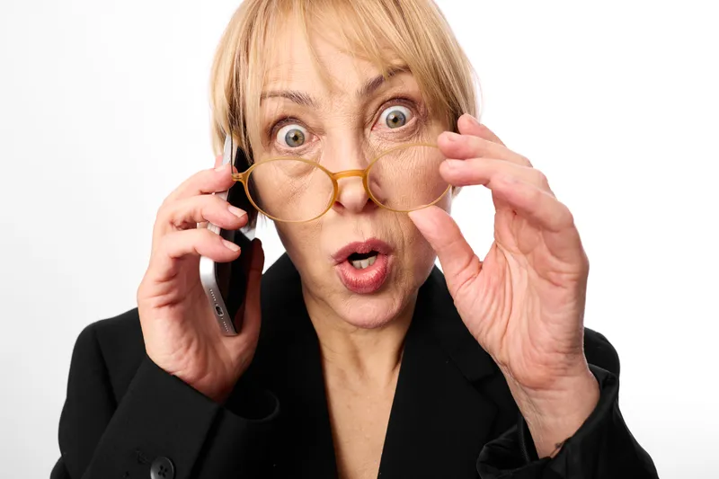 Woman in business suit speaks on phone while adjusting glasses with wide surprised eyes, hands near face, expressing stress and anxiety, communication concept on white background