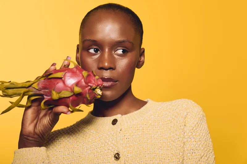 Portrait of a dark skinned African woman holding a dragon fruit near her face against a yellow background. Concept of healthy lifestyle, exotic fruit, and natural beauty.