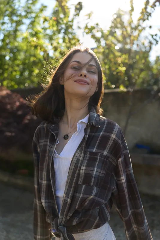 Confident woman outdoors in plaid shirt enjoys sunny day, casual lifestyle portrait with smiling expression, natural light, garden backdrop, relaxed mood and everyday fashion