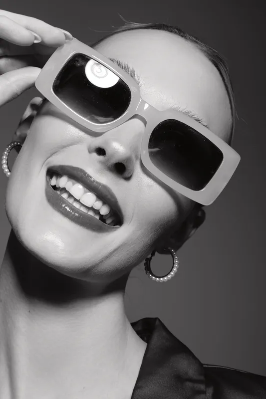 Smiling white woman with light skin wearing large sunglasses and hoop earrings in studio portrait with black background.