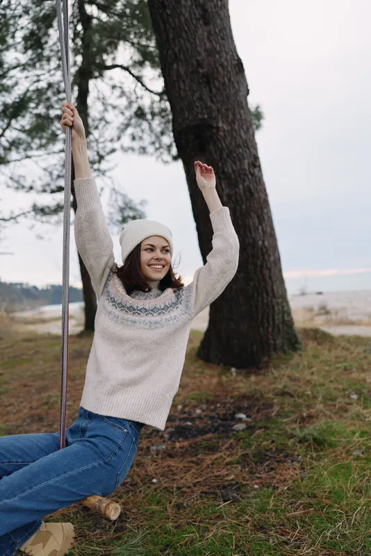 Girl on a swing in an outdoor park, wearing a cozy sweater and beanie, smiling while enjoying nature, trees and open space on a cool day with soft light and calm atmosphere.