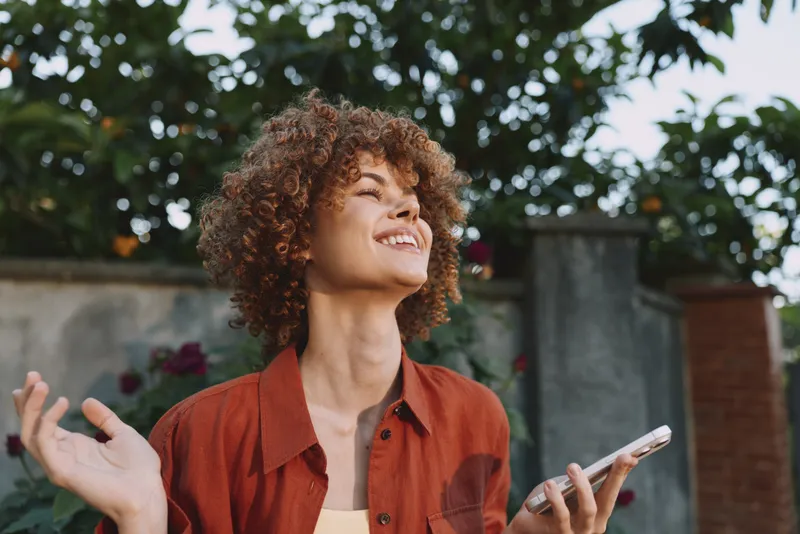 Smiling woman with curly hair in a red shirt outdoors, holding a phone. Natural light highlights lifestyle, wellness, and self care in a sunny garden setting