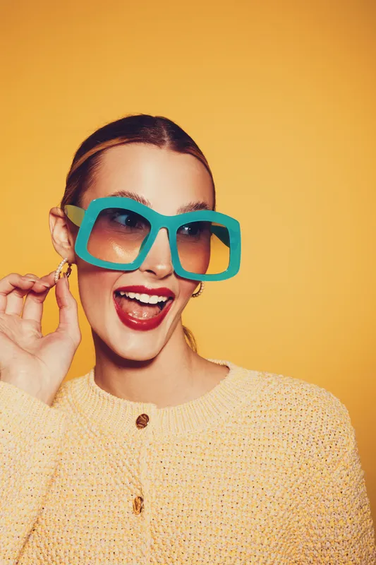 Happy woman with large blue sunglasses and red lipstick smiles brightly against yellow background. She wears a textured beige sweater and gold earrings. Concept of fashion and joy.