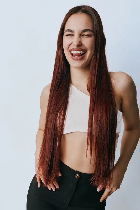 Young woman with long red hair and white top smiling and sticking out tongue in studio. Casual pose with hands in pockets on light background for lifestyle and happiness.