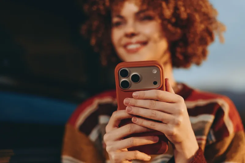 Smiling woman with curly hair holding smartphone for a selfie, candid woman lifestyle scene, warm indoor light, soft focus background, casual home setting, relaxed mood, close up portrait