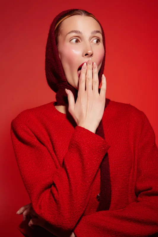 Surprised woman in red sweater and headscarf covers mouth with hand, looking sideways on red background. Concept of shock, emotion, and unexpected reaction in fashion portrait.