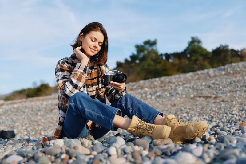 Woman sitting on a pebbled beach using a smartphone, wearing a plaid jacket and boots, relaxed in natural light with a calm, candid moment away from crowds and noise.
