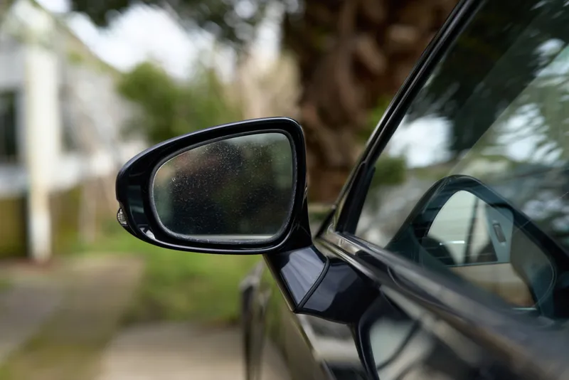 Close up of a car side mirror with raindrops, reflecting a blurred street and greenery, highlighting a sleek dark exterior and a moment of calm travel