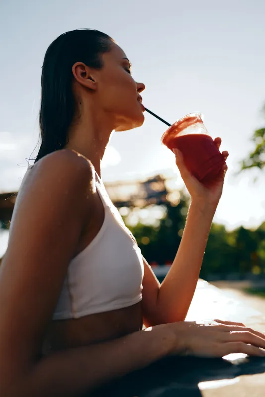 A young woman in a white bikini top sips a pink smoothie with a straw, relaxing by a sunny pool on a vibrant day, her eyes calm and the bright water glowing behind her.