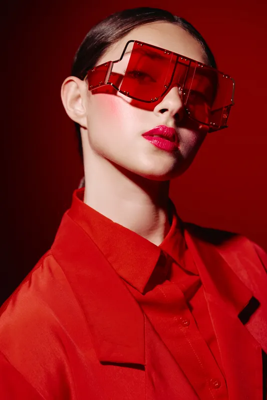 Fashion model wearing oversized red sunglasses and vibrant red suit jacket poses confidently against a red background. Makeup focused on intense glossy lips. Creative concept for beauty and style.