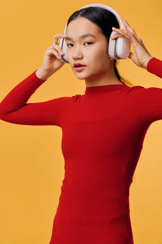 Young woman in red dress wearing white headphones listening to music with closed eyes on yellow background. Asian female enjoying sound, lifestyle and modern entertainment technology.