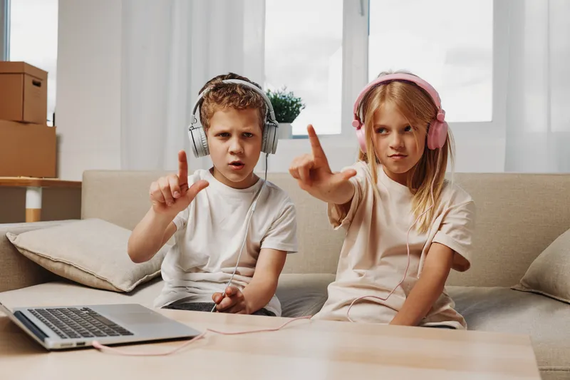 Two children wearing headphones sit on a sofa at home using a laptop, focused on listening and interacting with digital content in a casual living room setting.