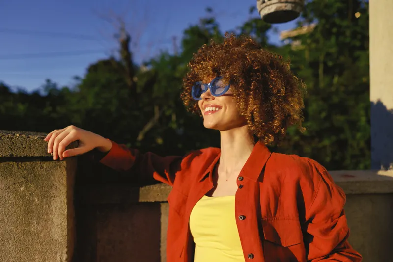 Radiant woman with curly hair wears an orange jacket and yellow top, posing outdoors in bright sun, expressing a cheerful lifestyle and confident fashion sense of style and joy