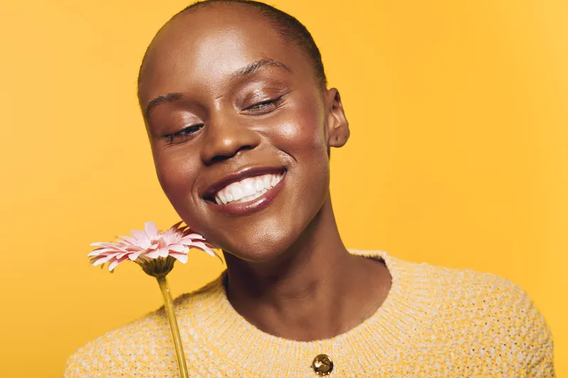 Smiling black woman with dark skin holds pink flower close to face, wearing yellow sweater. Studio portrait on yellow background. Concept of happiness, beauty, and natural skincare.