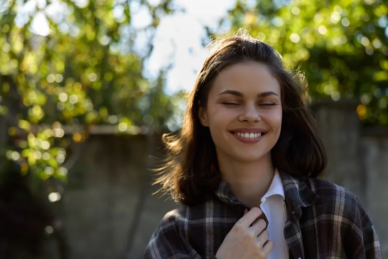 Smiling woman outdoors wearing a plaid shirt, close-up portrait on a sunlit background. Focus on lifestyle, fashion, and natural beauty
