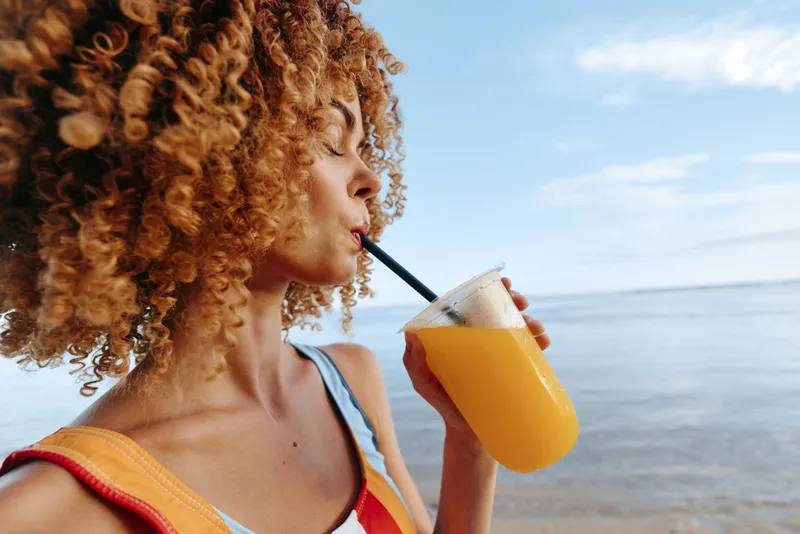 Curly woman drinks orange juice from plastic cup with straw at beach on sunny day, enjoys fresh tropical drink with sea and sky background, casual summer lifestyle concept
