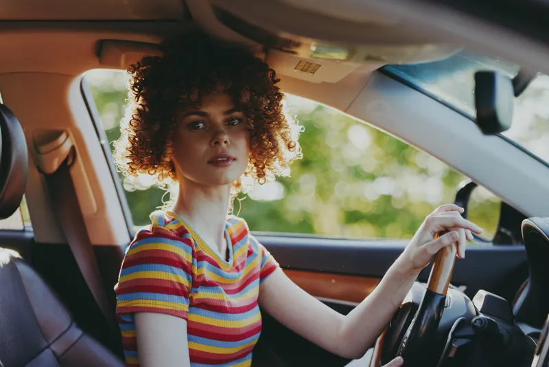 A young woman with curly hair sits in a car, wearing a striped tee, looking ahead with a thoughtful gaze as warm retro film tones wash over the scene.