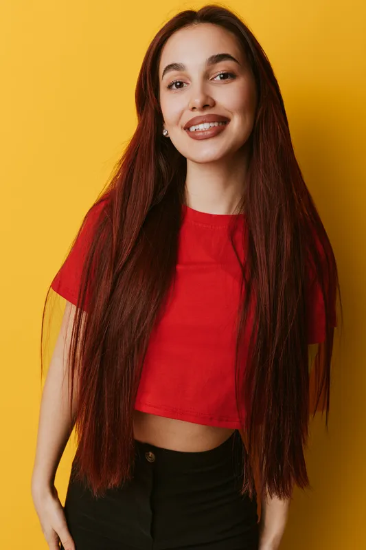 Young woman with long straight hair wearing red crop top and black pants smiling confidently against yellow background in studio portrait with casual style and natural makeup.