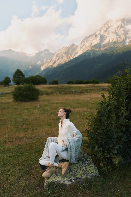 Woman in a serene mountain meadow lifestyle sits on a rock wearing a light sweater, enjoying nature and a calm outdoor moment with distant peaks and blue sky mood.