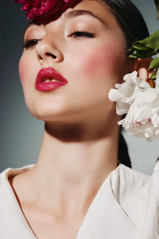 Close-up of a woman with bold pink lips and flushed cheeks wearing dramatic floral accessories in her hair. Studio portrait focused on beauty fashion and creative makeup.