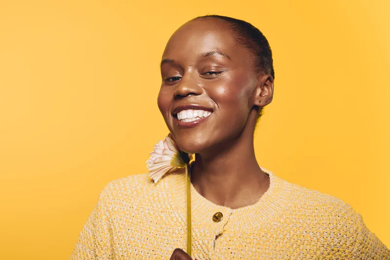 Happy African woman with dark skin smiling and holding a flower near her face. Yellow background and cozy sweater create warm and cheerful mood. Concept of beauty and joy.