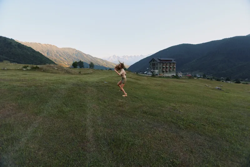 Young woman jumps joyfully on green grass field in mountain valley near house during sunny day, enjoying active lifestyle and nature outdoors.