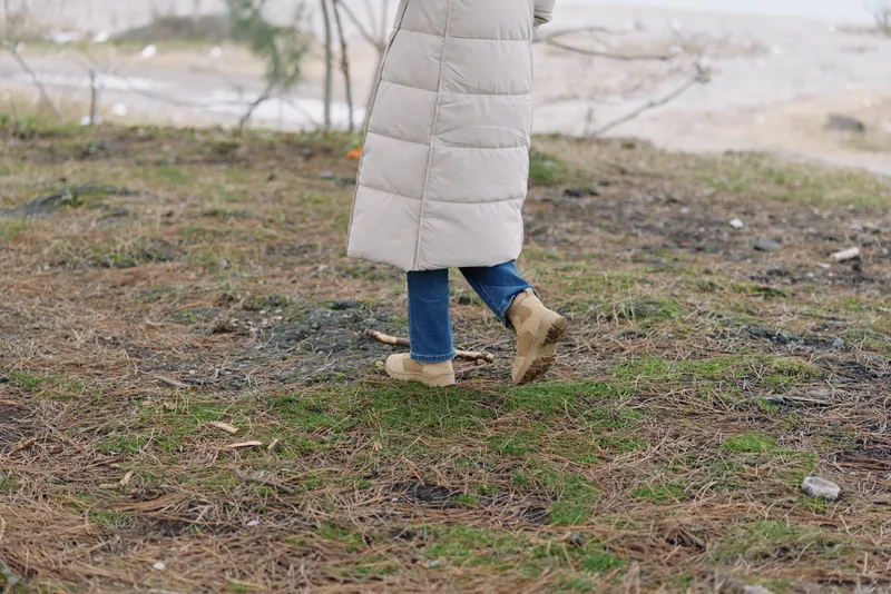 Person walking outdoors in a light quilted coat, blue jeans and sturdy boots along a natural path beside a sparse landscape