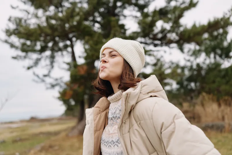 Outdoor portrait of a woman in a warm coat and beanie, standing in a windy landscape with trees and open fields. A calm, candid moment showing comfort in chilly weather and seasonal fashion