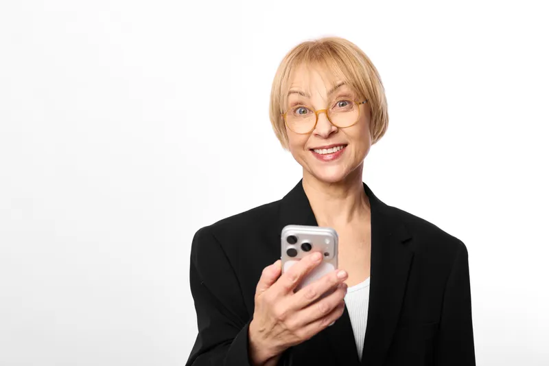 Senior woman with glasses smiling, holding a smartphone, studio portrait on white background, relaxed posture, friendly expression, modern communication