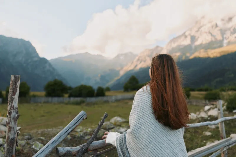 Woman with long hair in a knit sweater stands beside a wooden fence gazing at distant mountains, serene outdoor lifestyle scene, nature and wide open space.