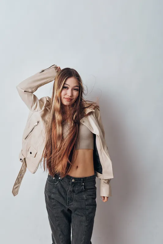 Young woman with long hair wearing a light jacket and crop top posing against white background in casual style with relaxed and confident expression.