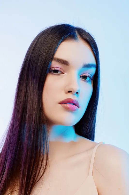 Close-up portrait of a young woman with long straight hair and flawless skin. Soft blue lighting highlights her beauty and fashion style in a minimal studio setting.