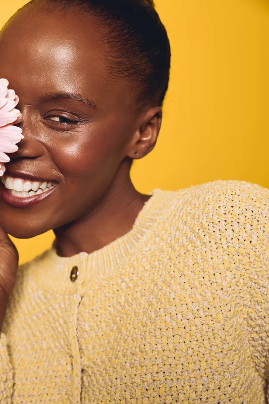 Smiling African woman with dark skin holds pink flower close to face. She wears a yellow knit sweater and stands against a bright yellow background. Concept of happiness, beauty, and natural skin.