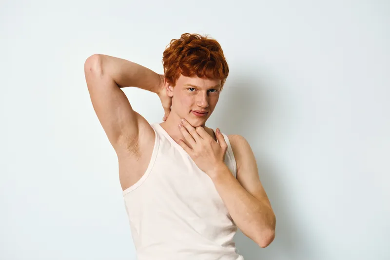 Young man with red hair wearing white tank top posing with hand behind head and touching neck against plain white background for casual portrait.