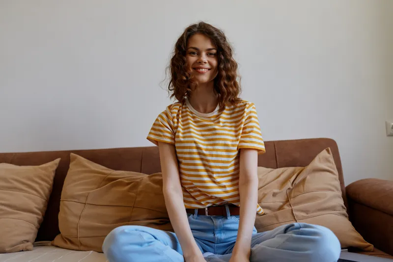 Young woman sits on a cozy sofa at home, wearing a striped t shirt and blue jeans, smiling softly as warm natural light pours into the bright living room