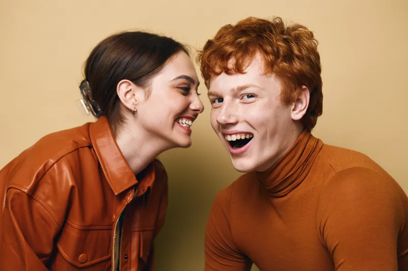 Young man and woman laughing and talking closely in casual autumn clothing with bright expressions against plain background. Friendship and joy captured in studio portrait.