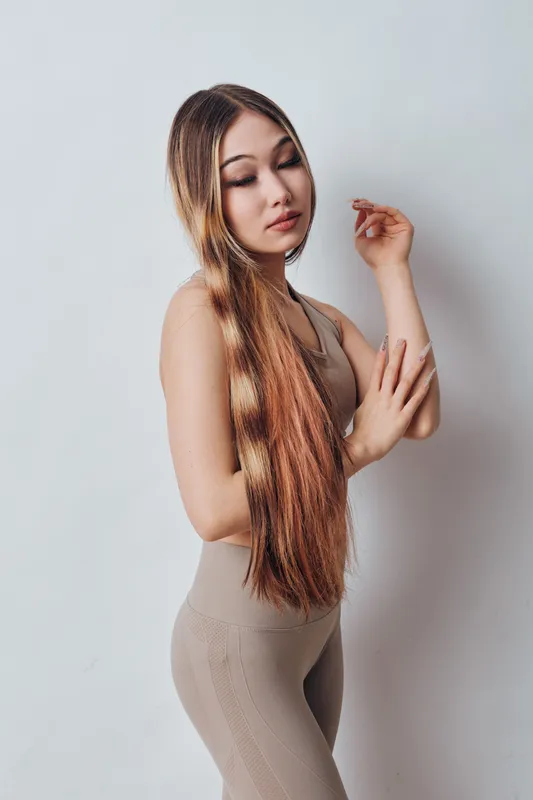 Young woman with long hair posing in a sleeveless beige outfit standing against a plain light background. Beauty, hairstyle and fashion portrait in studio setting.