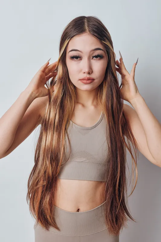 Young woman with long straight hair standing against white background wearing casual beige crop top and matching pants, looking at camera with relaxed expression.