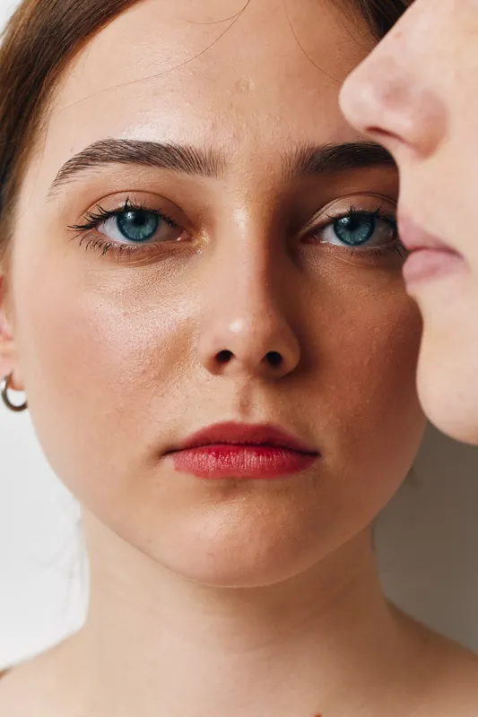Portrait of young woman with blue eyes and clear skin looking forward indoors with profile of another person close by for facial expression and emotion concept.