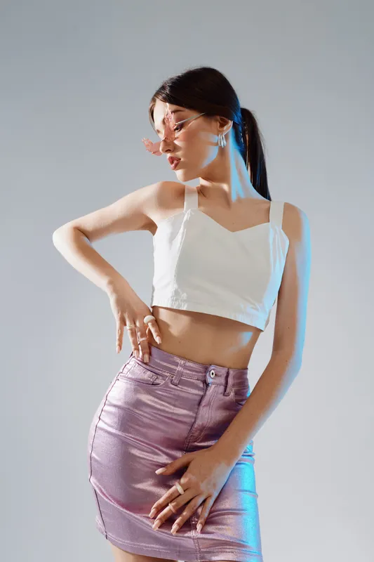 Young woman in a white crop top and shiny skirt poses confidently with sunglasses. Studio shot with soft lighting. Concept of woman beauty fashion and style.
