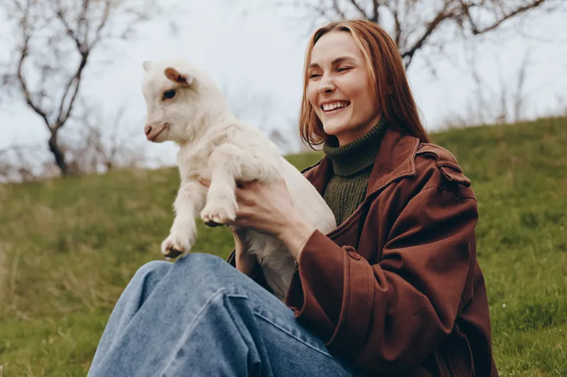 Young woman with long red hair smiles happily while holding a small white baby goat outdoors on a grassy hill in casual autumn clothing.