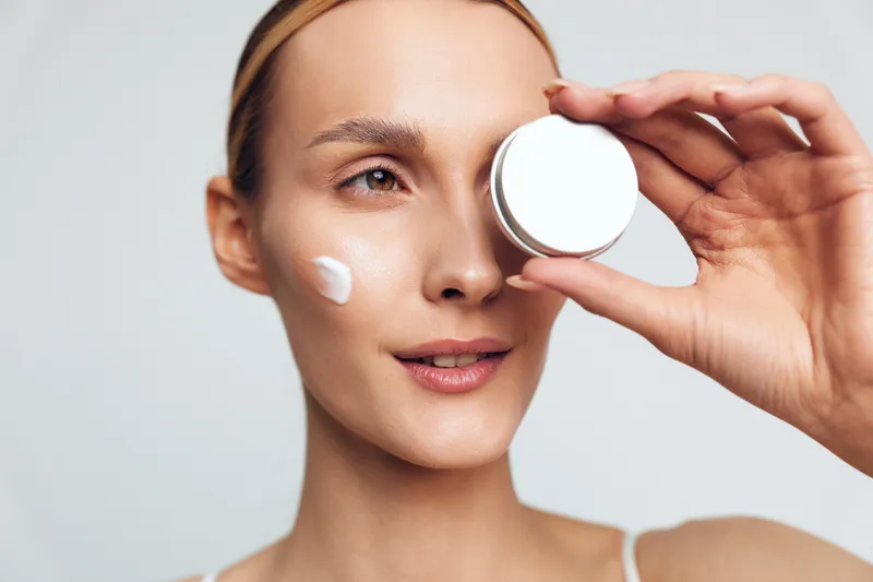 Young woman with clear skin holding a white cream jar near her eye, applying face cream on cheek. Concept of skincare, beauty routine, wellness, and self-care with soft lighting.