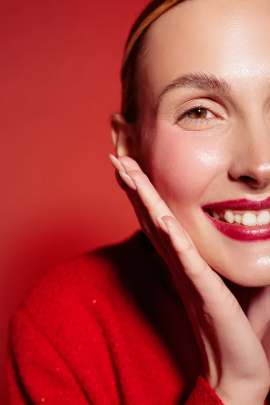 Close-up of smiling woman with red lipstick and red sweater touching her face. Bright red background and natural skin glow create warm and cheerful mood. Concept of beauty and happiness.